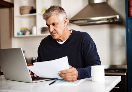 Middle-aged man reading on laptop at kitchen table with papers in his left hand.